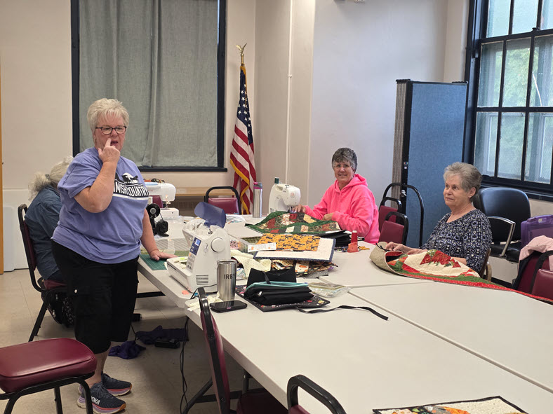 A member standing and chatting while others sew and lay out quilt pieces