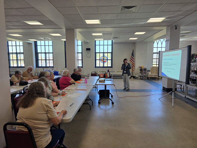 A presenter speaking to members seated at tables in the bright main hall
