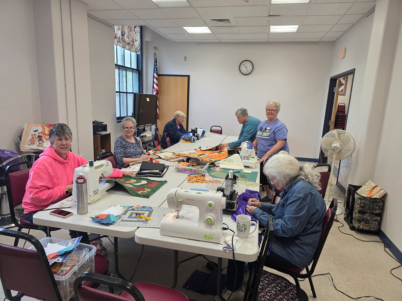 Members working together at sewing machines on colorful quilts around a long table