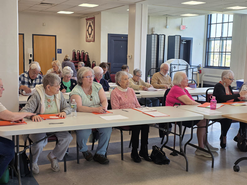 Members seated at tables with handouts, listening to a presentation